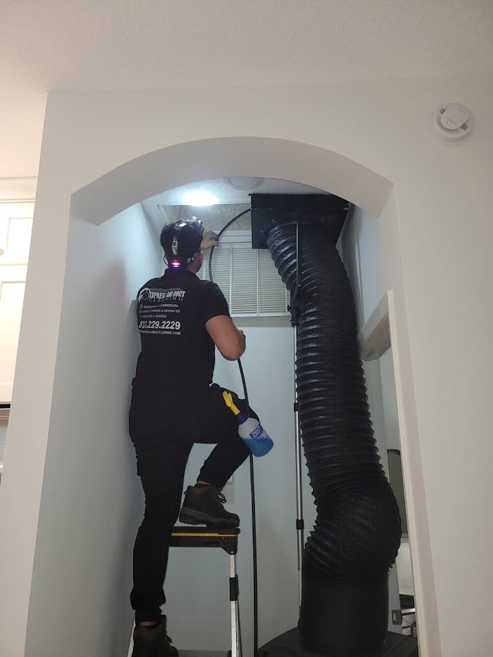 Express Air Duct Cleaning technician on a ladder cleaning a ceiling air duct with a black flexible hose